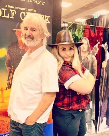 A smiling picture of John Jarratt in white shirt with his co-actress on shooting day 